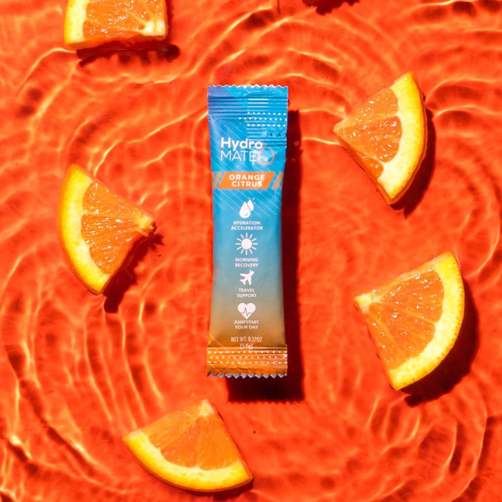 Single-serving packet of Hydro Mate Orange Citrus hydration accelerator floats on a vibrant orange, water-like background with fresh orange slices. 
